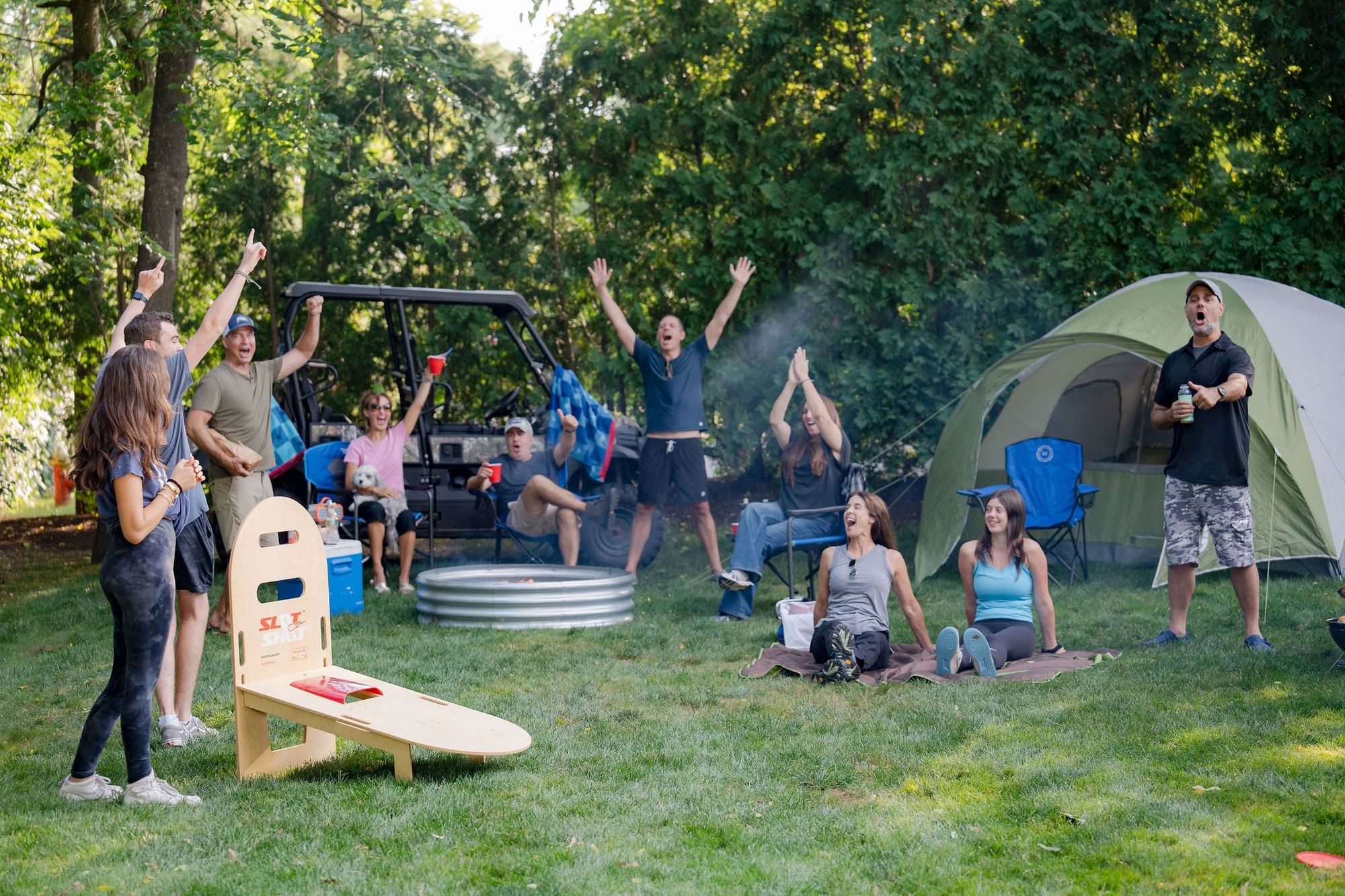 Group of friends cheering while playing SlotShot backyard disc game near a tent and camp setup during summer
