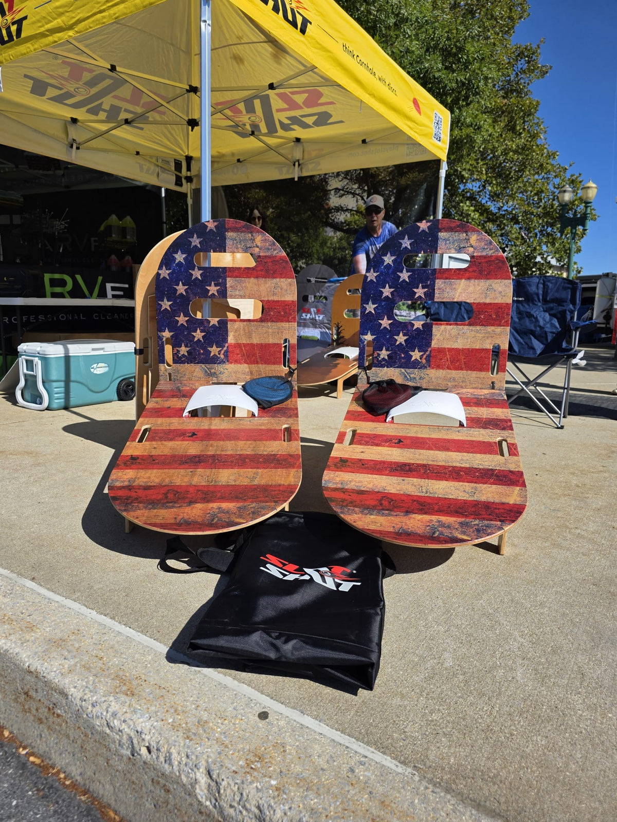 Two wooden SlotShot boards with a distressed American flag design,, set outdoors under a yellow tent with a black bag in front on a concrete surface.