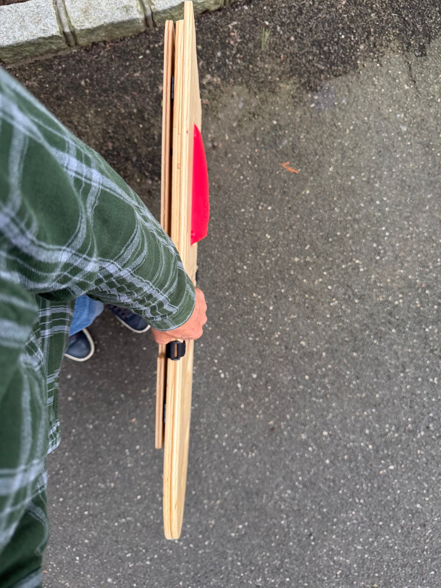 A person wearing a green plaid flannel shirt holds a long, narrow wooden board or collapsed SlotShot board with a bright red scoop and a small black circular detail which is a fastener to keep the boards together, standing on an asphalt surface outdoors.