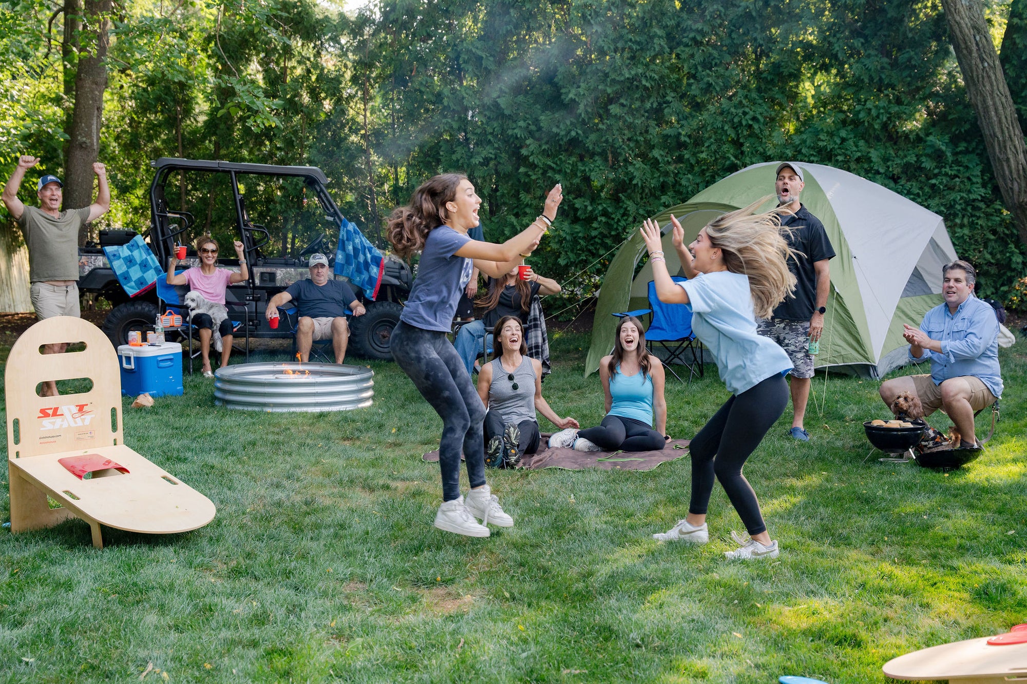 Group of friends celebrating while playing SlotShot backyard disc game at a campsite with tent and fire pit in background