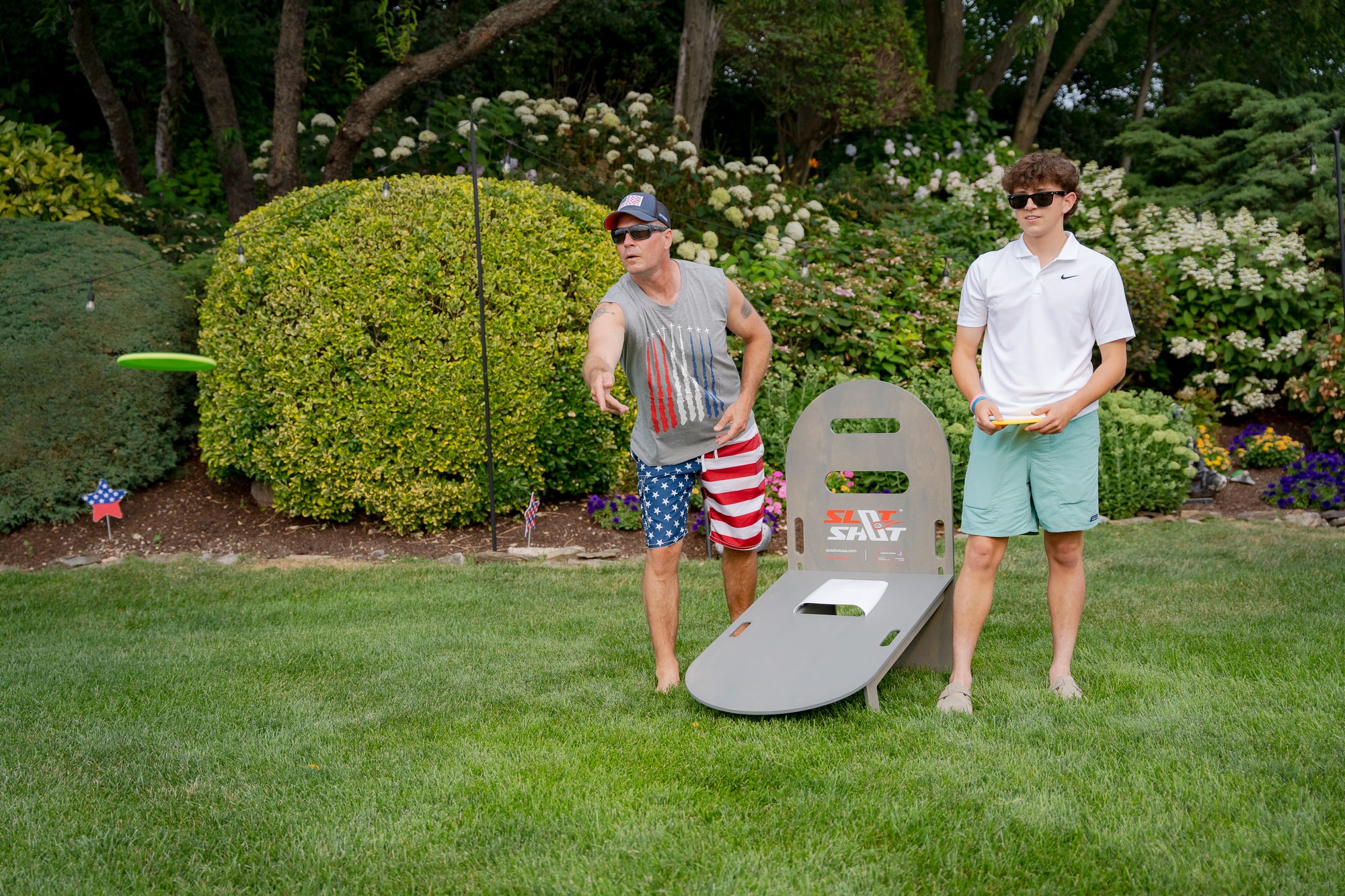Two people playing SlotShot backyard disc game on the lawn, one throwing a green disc wearing American flag shorts and tank top