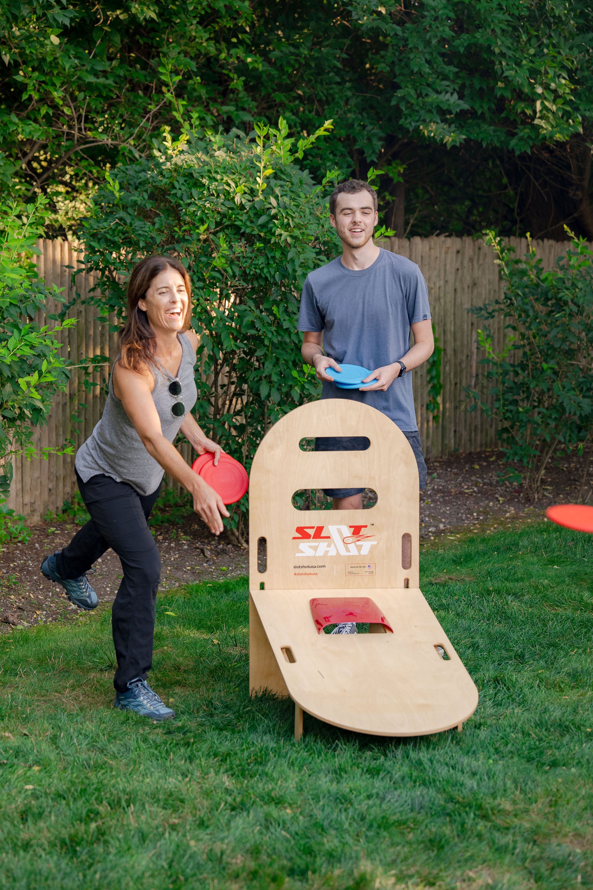 Two friends playing SlotShot backyard disc game on the grass with red and blue discs during a sunny afternoon
